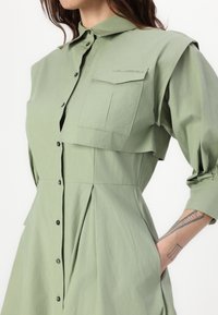 Rochie verde deschis, cu guler și butoni, având o buzunar pe piept, un design cu fustă plisată și o textură a materialului care pare ușoară și netedă.