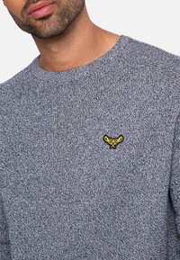 Grauer Strickpullover mit Rundhalsausschnitt und strukturiertem Gewebe. Enthält ein kleines gelbes Logo-Detail auf der Brust.