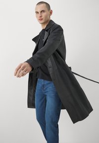 Schwarzer Leder-Trenchcoat mit spitzem Kragen, ausgestattet mit einem taillierten Gürtel und Klappentaschen, getragen über dunkler Kleidung und blauen Jeans.