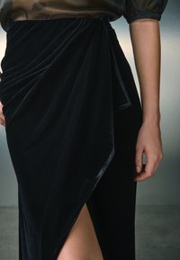Black velvet skirt featuring a draped design with pleats and a high slit. The fabric has a soft texture, enhancing its elegant appearance.