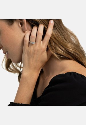 Verlobungsring mit einem runden Mittelstein, in einem eleganten Metallband gefasst, getragen an einer Hand mit natürlichen Nägeln, vor einem neutralen Hintergrund.