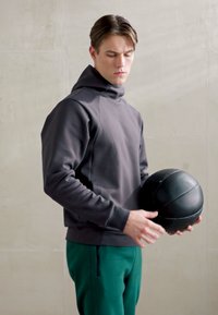 Felpa grigia con cappuccio dal tessuto morbido, abbinata a pantaloni della tuta verdi; tiene in mano un pallone da basket nero in un ambiente neutro.