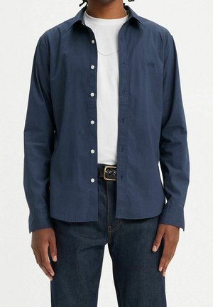 Muž v tmavě modré košili na knoflíky přes bílé tričko, tmavých džínách a černém pleteném opasku se zlatou přezkou, stojící před jednobarevným pozadím.