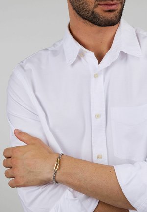 Braccialetto a catena d'argento con una chiusura in oro, indossato al polso. La persona indossa una camicia bianca con bottoni, le braccia incrociate, mostrando i dettagli del braccialetto.