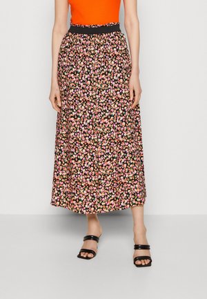 Kvinde iført sort blomsterprint midi nederdel, orange ærmeløs top og sorte åbne sandaler står mod en hvid baggrund.