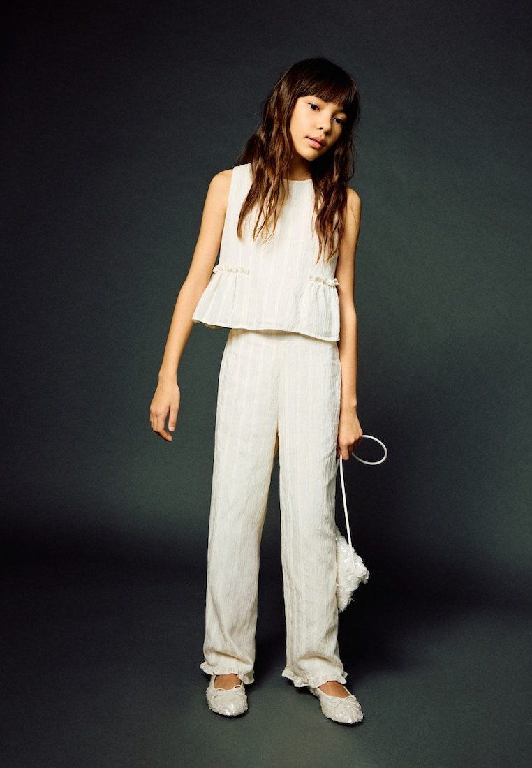 Jeune fille vêtue d'un haut blanc sans manches et d'un pantalon, tenant un petit sac à main blanc texturé sur un fond sombre.