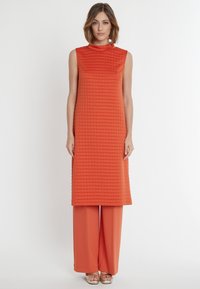 Orange ärmellose Tunika mit strukturiertem quadratischem Muster, kombiniert mit passenden weiten Hosen, abgerundet mit metallischen Sandalen.