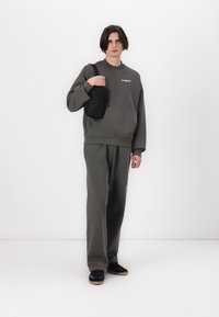 Pilkas sportinis megztinis su logo, derančios pilkos sportinės kelnės ir juoda per petį nešama rankinė. Modelis dėvi juodus sportinius batus ir stovi ant balto fono.