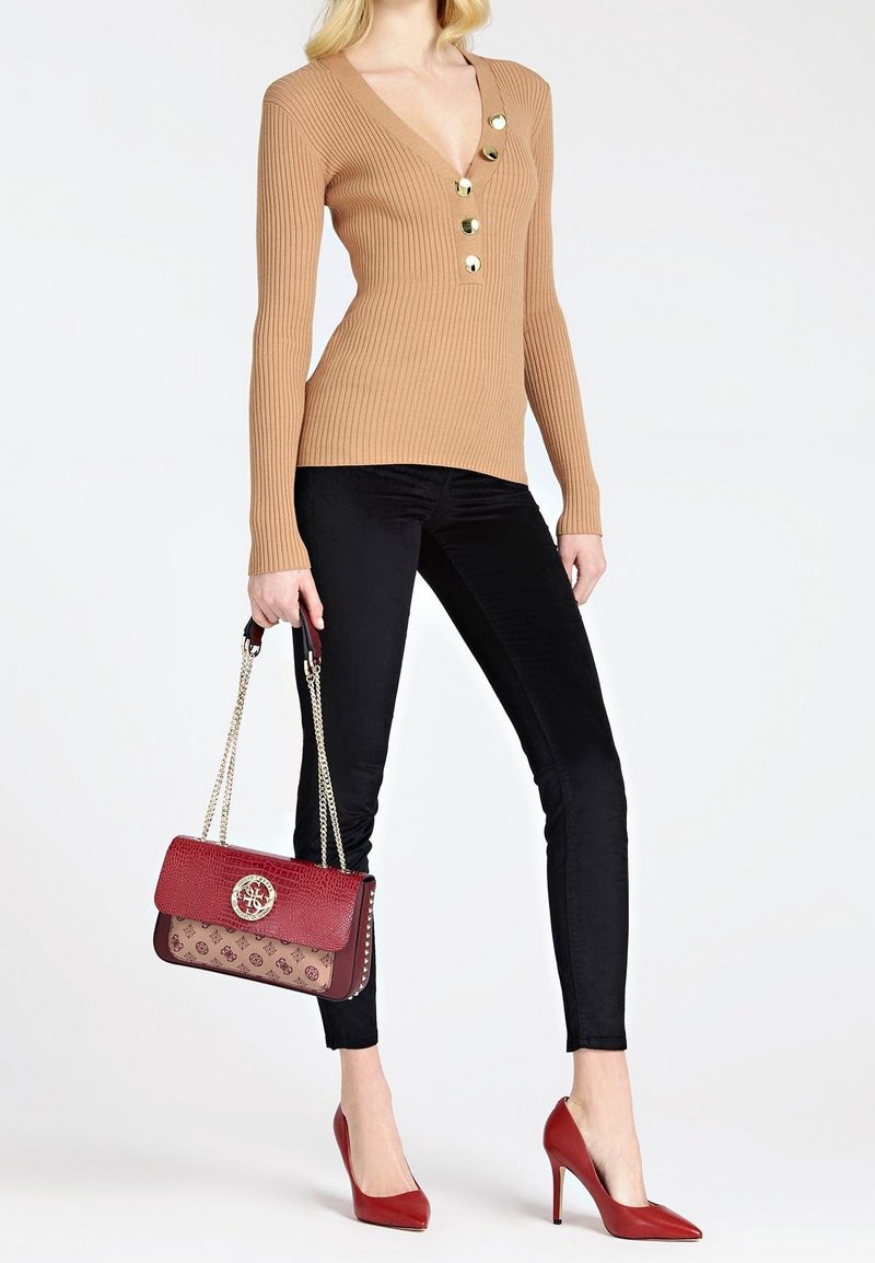 Femme portant un pull côtelé beige à col en V avec des boutons dorés, un pantalon skinny noir, des talons hauts rouges, et tenant un sac à main marron et rouge avec des anses chaîne.