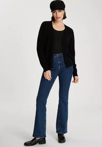 Czarny kardigan o luźnym kroju, noszony na dopasowanej czarnej bluzce, w połączeniu z dżinsami w stylu dzwonów w kolorze niebieskim i czarnymi mokasynami. Modelka ma na sobie czarną czapkę.