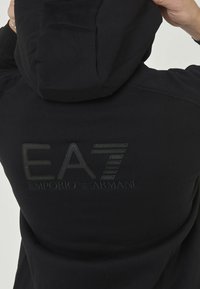 Sweatshirt noir à capuche avec logo brodé "EA7 Emporio Armani" de couleur assortie au dos, tissu lisse et capuche ajustable.