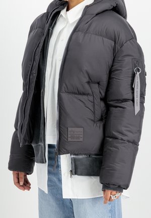 Osoba ubrana w czarną puchową kurtkę z kapturem, z zawieszką "Remove Before Flight" na rękawie, białą koszulę i jasnoniebieskie dżinsy.