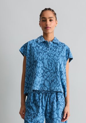 Vrouw die een blauw patroon kortärmelig top met kraag en bijpassende short met trekkoord draagt, staand tegen een lichtgrijze achtergrond.