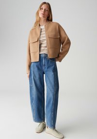 Giacca corta color talpa con due tasche anteriori, abbinata a un maglione a righe e jeans blu larghi. La modella indossa sneakers grigio chiaro.