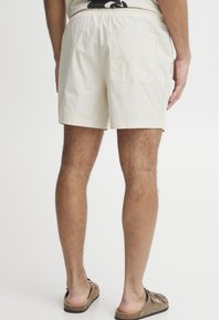 Pantalones cortos de algodón beige con cintura elástica, bolsillo trasero y corte relajado. Llevados con sandalias marrones que presentan correas texturizadas.