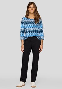 Blaues Oberteil mit Chevron-Muster und drei Viertelärmeln, kombiniert mit schwarzen Hosen. Das Model trägt weiße Sneakers. Einfaches Design, lässige Passform.