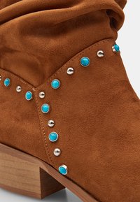 Botín de ante marrón con caña doblada, tachuelas plateadas y detalles en turquesa. Cuenta con un tacón de bloque de madera.