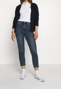Dunkelblauer Strickcardigan über einem weißen Poloshirt, kombiniert mit hochgeschnittenen, schmalen Jeans und weißen High-Top-Sneakern.