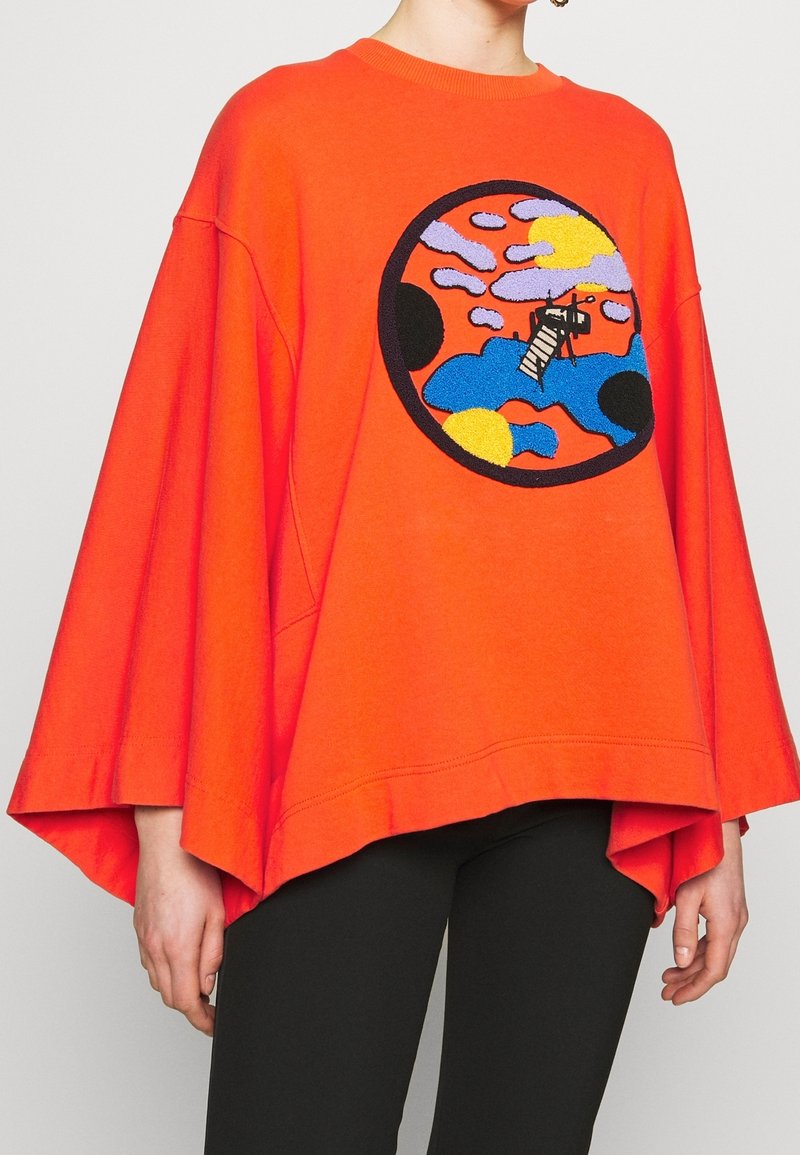 Pull ample orange vif à manches larges, arborant un motif abstrait circulaire coloré avec une échelle noire au centre.