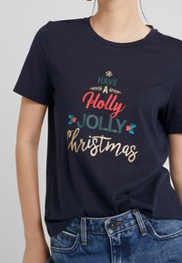 Laivastonsininen puuvillateepaita, jossa on juhlavasti tulostettu kuvio, jossa lukee "Have a Holly Jolly Christmas" vihreillä, punaisilla ja kultaisilla yksityiskohdilla.