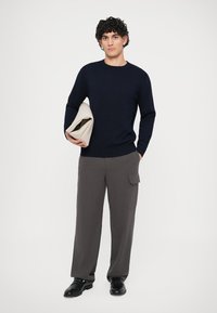 Ναυτικό πλεκτό πουλόβερ, χαλαρή εφαρμογή, υφή υφάσματος; γκρι παντελόνι σε φαρδύ κόψιμο με πλαϊνές τσέπες, φορεμένο με μαύρα παπούτσια, κρατώντας έναν μπεζ φάκελο.