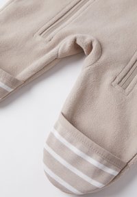 Beige Fleece-Overall mit zwei Reißverschlusstaschen. Die Ärmel und Beine haben gestreifte Bündchen in hellbeige und weiß. Weicher Stoff, warmes Design.