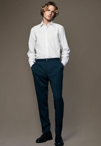 White long-sleeve shirt with a collar, paired with dark blue trousers featuring a button closure. Black leather shoes complete the outfit.