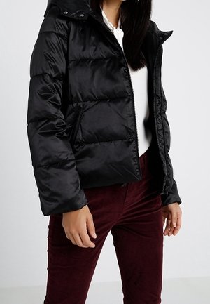 Vrouw draagt een zwarte pufferjack over een witte top en donkerrode broek, staand met ontspannen handen langs het lichaam tegen een effen achtergrond.