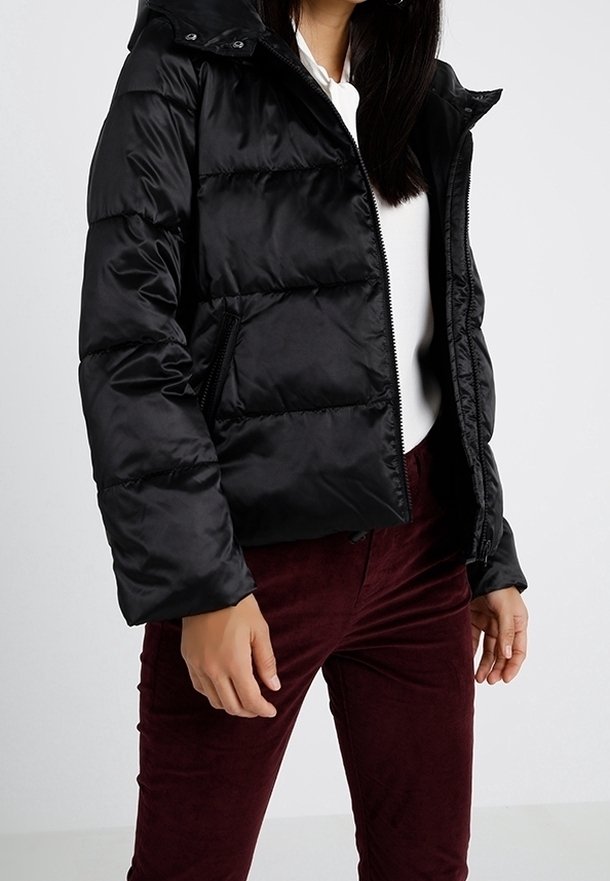 Femme portant une doudoune noire sur un haut blanc et un pantalon rouge foncé, debout les mains détendues le long du corps devant un fond uni.