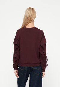 Γυναίκα με μακριά ξαν�θά μαλλιά φοράει ένα σκούρο μωβ πουλόβερ με στρώσεις διαφανούς φουσκωτού μανικιού και σκούρα μπλε τζιν, στέκεται με την πλάτη στραμμένη.