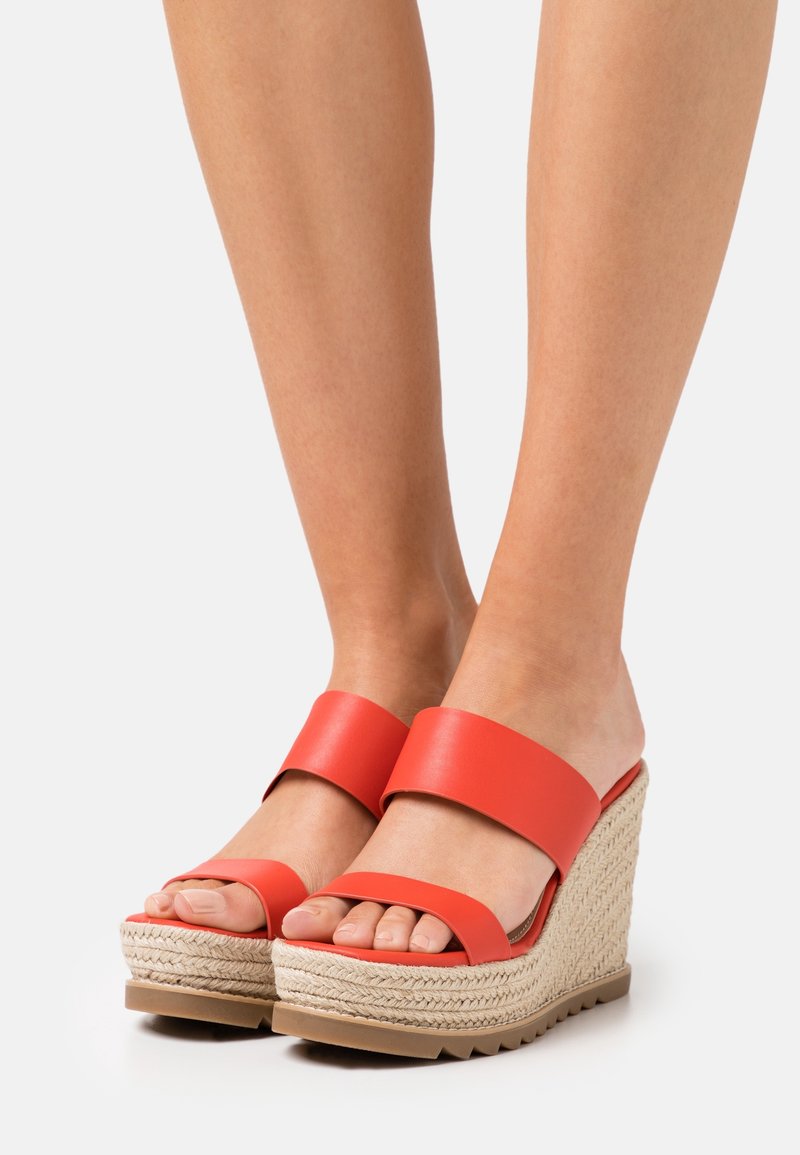 Pieds portant des sandales compensées rouges à bout ouvert avec des semelles plateformes en paille tressée sur un fond blanc uni.