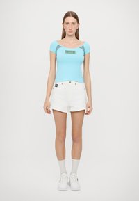 Light blue fitted short-sleeve top with a logo patch, paired with white high-waisted shorts. Model wearing white sneakers and socks.