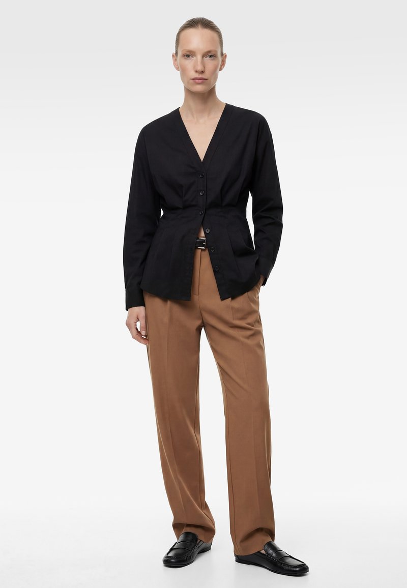 Kobieta stoi ubrana w czarną, zapinaną na guziki bluzkę z dekoltem w serek, brązowe spodnie z wysokim stanem z paskiem oraz czarne mokasyny na białym tle.