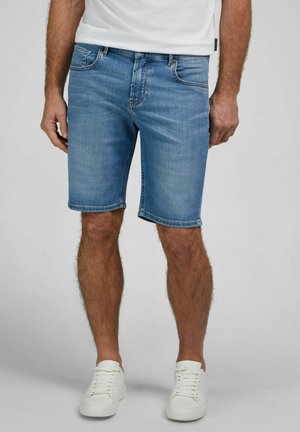 Mann trägt hellblaue Jeansshorts, weiße Turnschuhe und ein weißes T-Shirt, steht vor einem einfarbigen grauen Hintergrund.