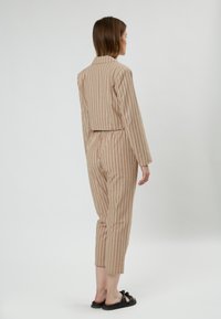 Jumpsuit a strisce beige con maniche lunghe, design cropped, vita elastica e pantaloni tapered alla caviglia. Indossato con sandali neri.