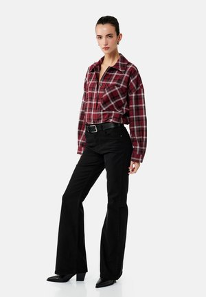 Kvinde iført en rød ternet skjorte med knapper, tucked ind i højtaljede sorte vidde bukser med sort bælte og sorte hælede støvler.