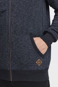 Navy-blue zip-up hoodie with a textured fabric, featuring front pockets and a brown logo patch. Cuffs and hem are solid navy.