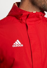 Gros plan d'une personne portant une veste rouge Adidas avec capuche, montrant la partie supérieure de la poitrine et du cou.