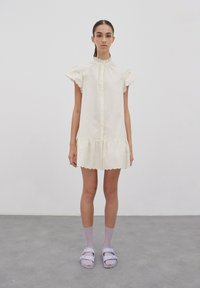 Weißes Baumwollkleid mit gewellten Kanten, kurzen Rüschensäumen, Knopfverschluss vorne und gestuften Saum. Getragen mit lila Socken und Sandalen.