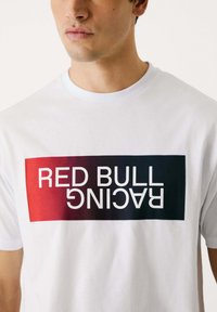 Balta īsām piedurknēm T-kreklā ar centrētu grafiku, kurā ir teksts "RED BULL RACING" ar sarkanā uz tumši zilā gradientu.