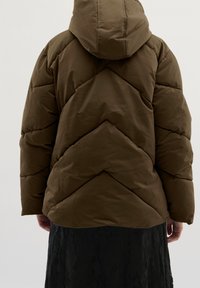 Brązowa kurtka puchowa z kapturem i wzorem w zygzak. Gładka faktura i luźny krój, noszona na czarnej spódnicy o fakturze.