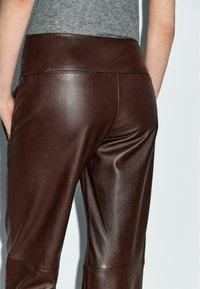 Persona che indossa pantaloni in pelle marrone scuro a vita alta e una maglietta grigia, con una mano parzialmente in tasca, su uno sfondo chiaro e semplice.
