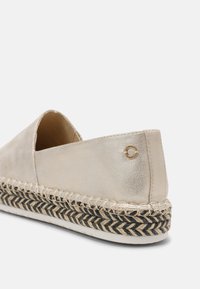 Espadrille beige avec un éclat métallique, dotée d'une semelle en jute tissée, d'accents noirs et d'un petit logo décoratif sur le talon.