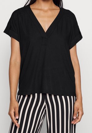 Kobieta ubrana w luźną czarną bluzkę z krótkim rękawem i dekoltem w serek, z podwiniętymi rękawami, oraz czarno-białe pionowo paskowane spodnie.