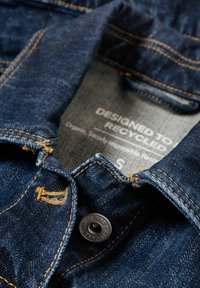 Dark blue denim jacket with visible stitching, a black metal button, and a label stating “Designed to Recycled” on the inside collar.