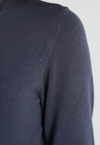 Pull en maille bleu foncé avec une texture côtelée et un col rond ; gros plan sur la couture de l'épaule et les détails du tissu.