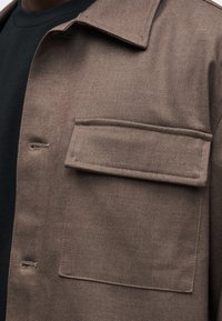 Κοντινό πλάνο σε τσέπη καφέ μπουφάν με καπάκι, εμφανείς οι τρύπες για κουμπιά, που φοριέται πάνω από μαύρο πουκάμισο.