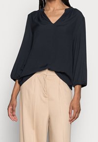 Blouse bleu marine foncé à col en V avec des manches 3/4 et des détails froncés au niveau du col. Le tissu a une texture lisse, associé à un pantalon large beige.