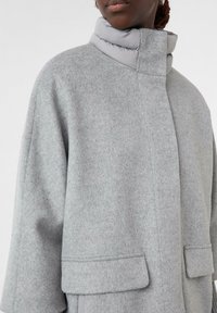 Grauer Wollmantel mit hohem, gepolstertem Kragen; verfügt über zwei Vordertaschen und eine lässige Silhouette. Strukturierter, weicher Stoff mit dezentem Muster.