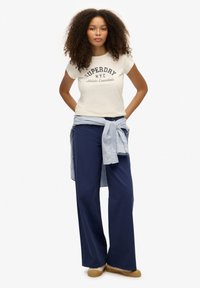 Camiseta crema con el texto "SUPERDRY N.Y.C.", combinada con pantalones de pierna ancha en navy. Una camisa de rayas azul claro está atada alrededor de la cintura. Zapatos marrones tipo slip-on.